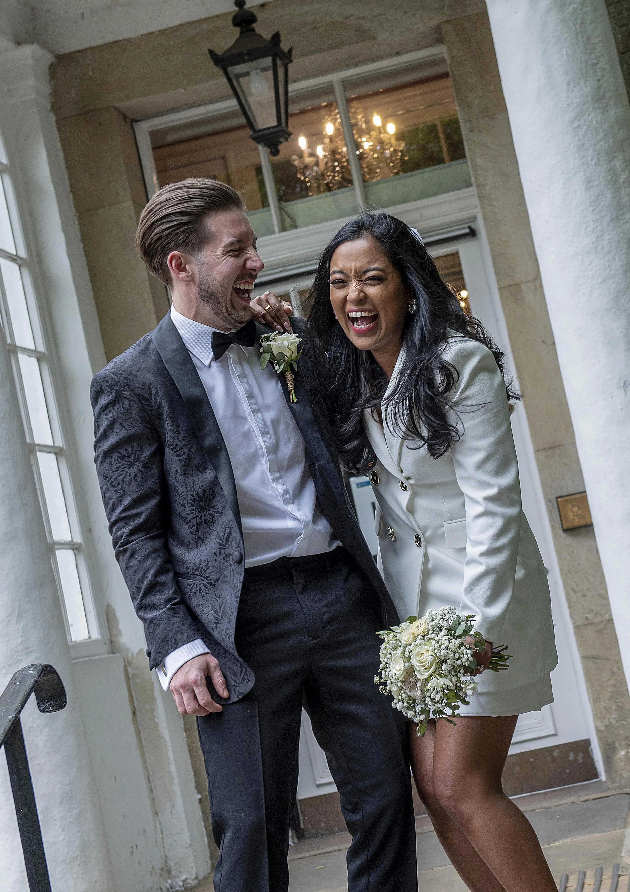 A man and woman sharing a joyful moment, the woman holding a bouquet of flowers, standing in front of a house entrance with a lantern hanging above. Harrogate wedding