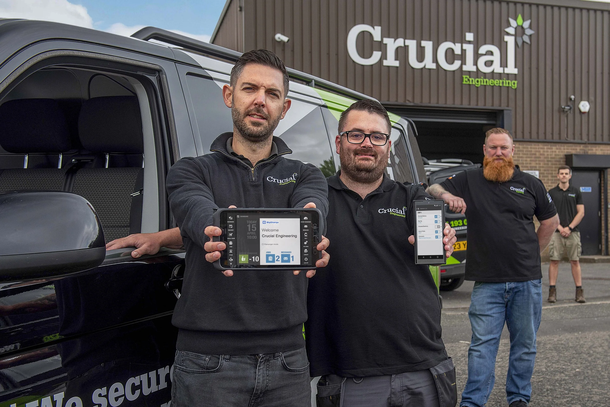 Three men standing outside a Crucial Engineering building, holding up devices showing digital screens, with a black vehicle behind them. The men are wearing black shirts with the company logo, and there are other people in the background.