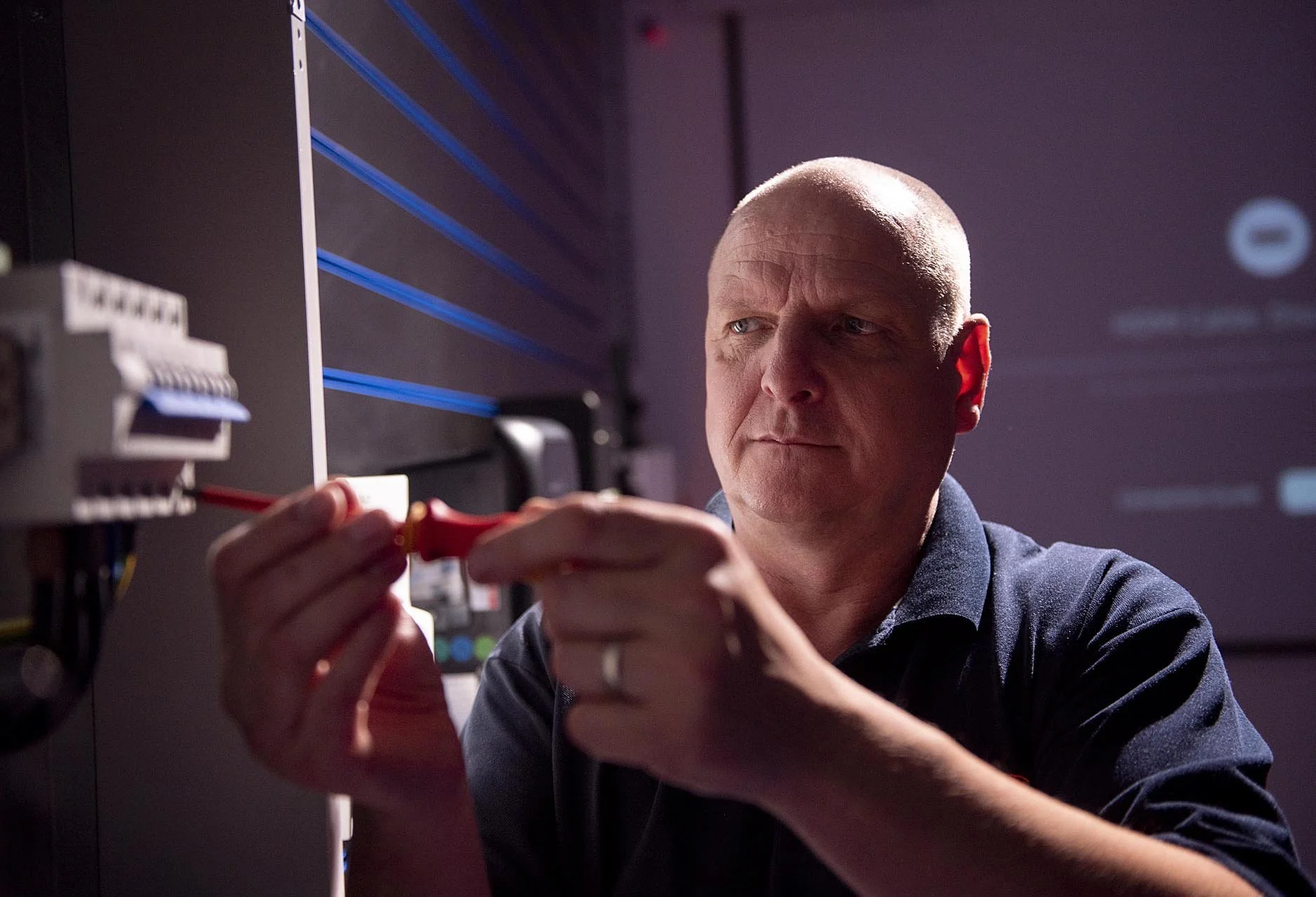 A man with a serious expression working with electrical wiring or equipment, using a screwdriver.