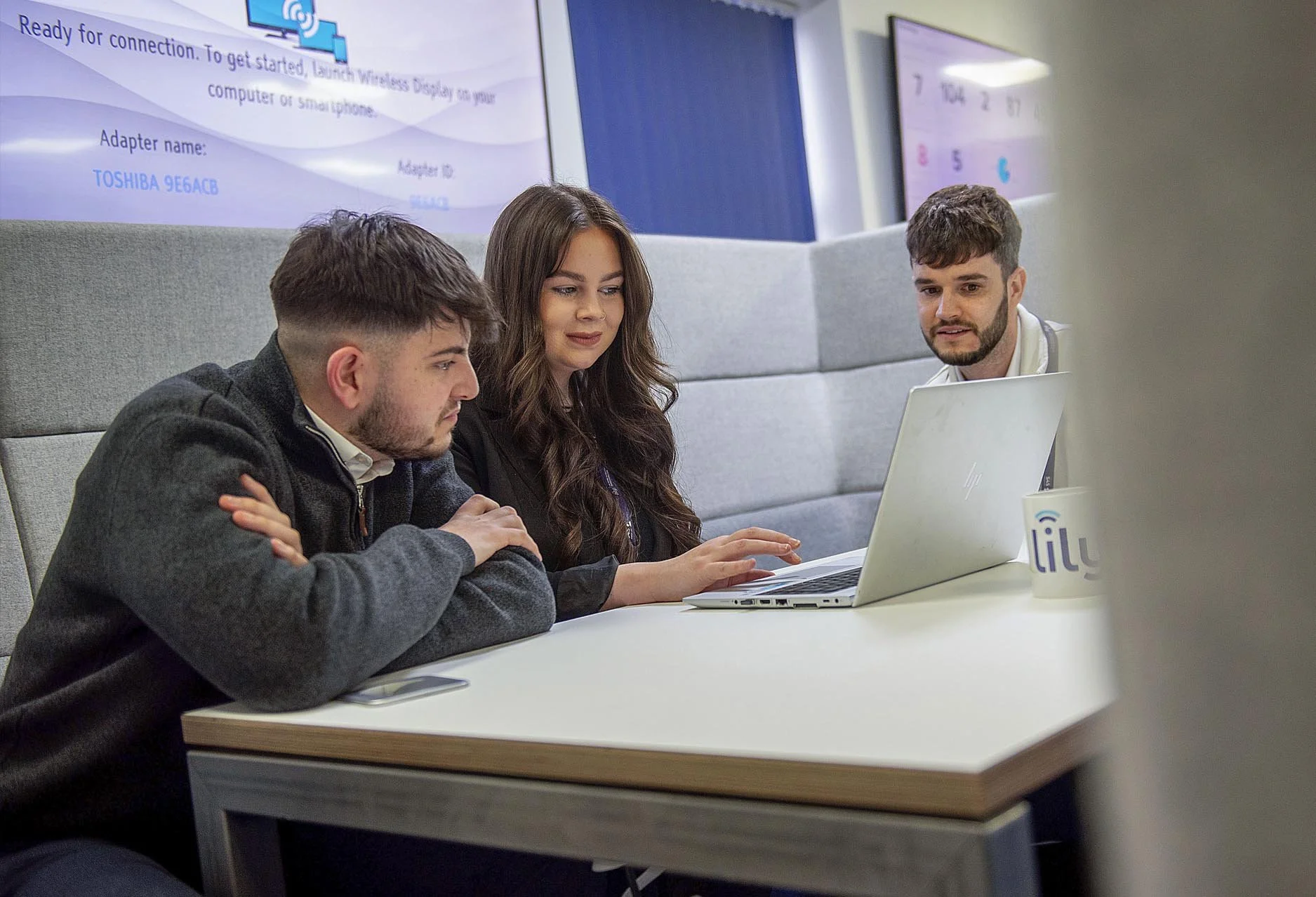 Three young adults sitting at a conference table working together on a laptop in a modern office space, with multiple screens and a Lily Wi-Fi device on the table.
