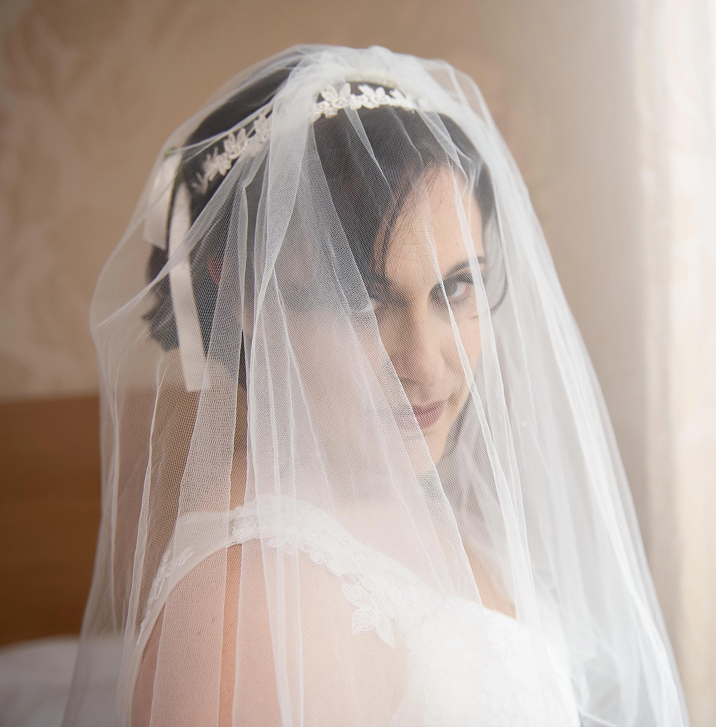 bride looking into camera through her veil standing next to a window. Leeds wedding