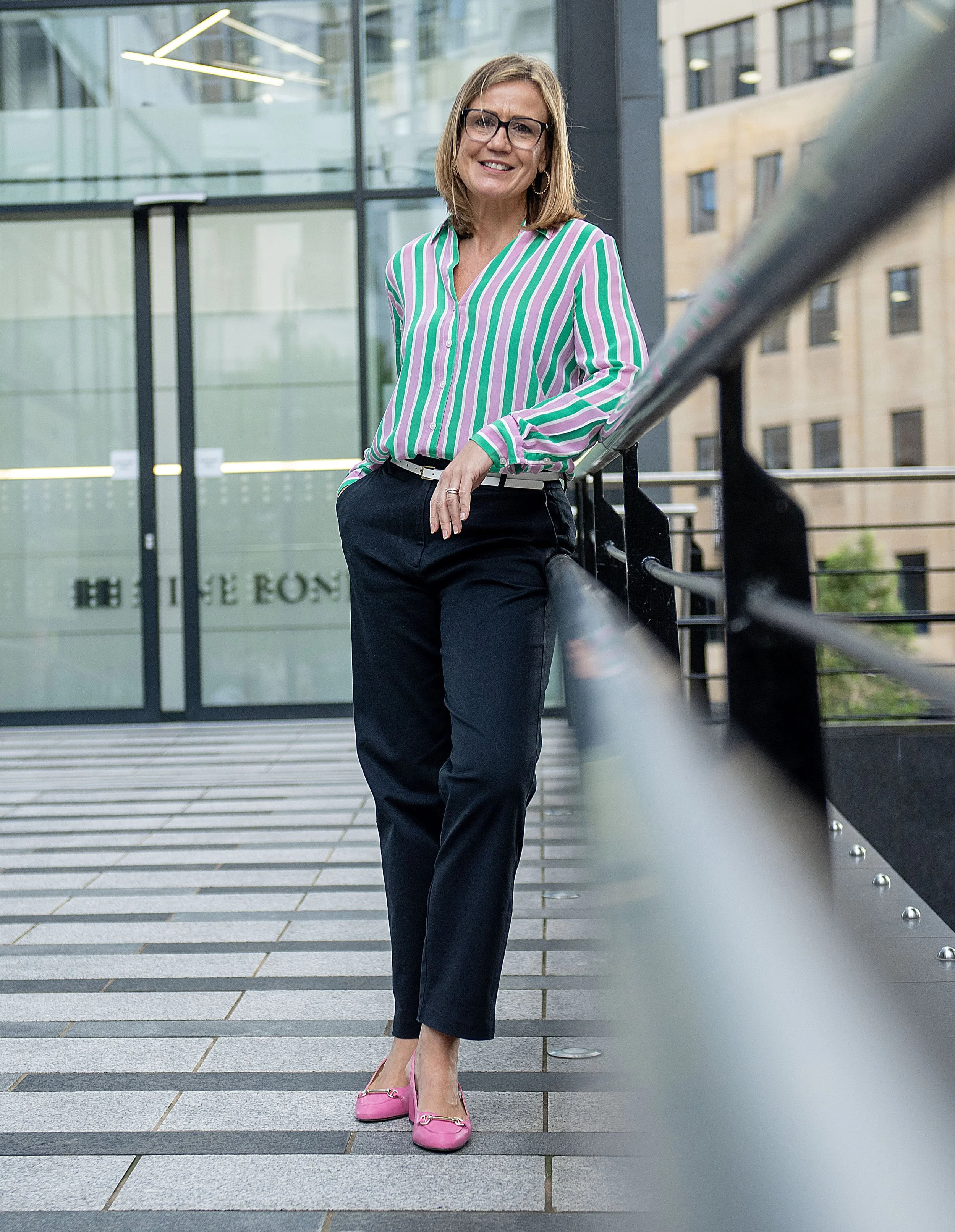 Woman in a teal striped shirt leaning on a railing outdoors, lifestyle business portrait against a glass office building. Corporate business photography session in Leeds city centre.