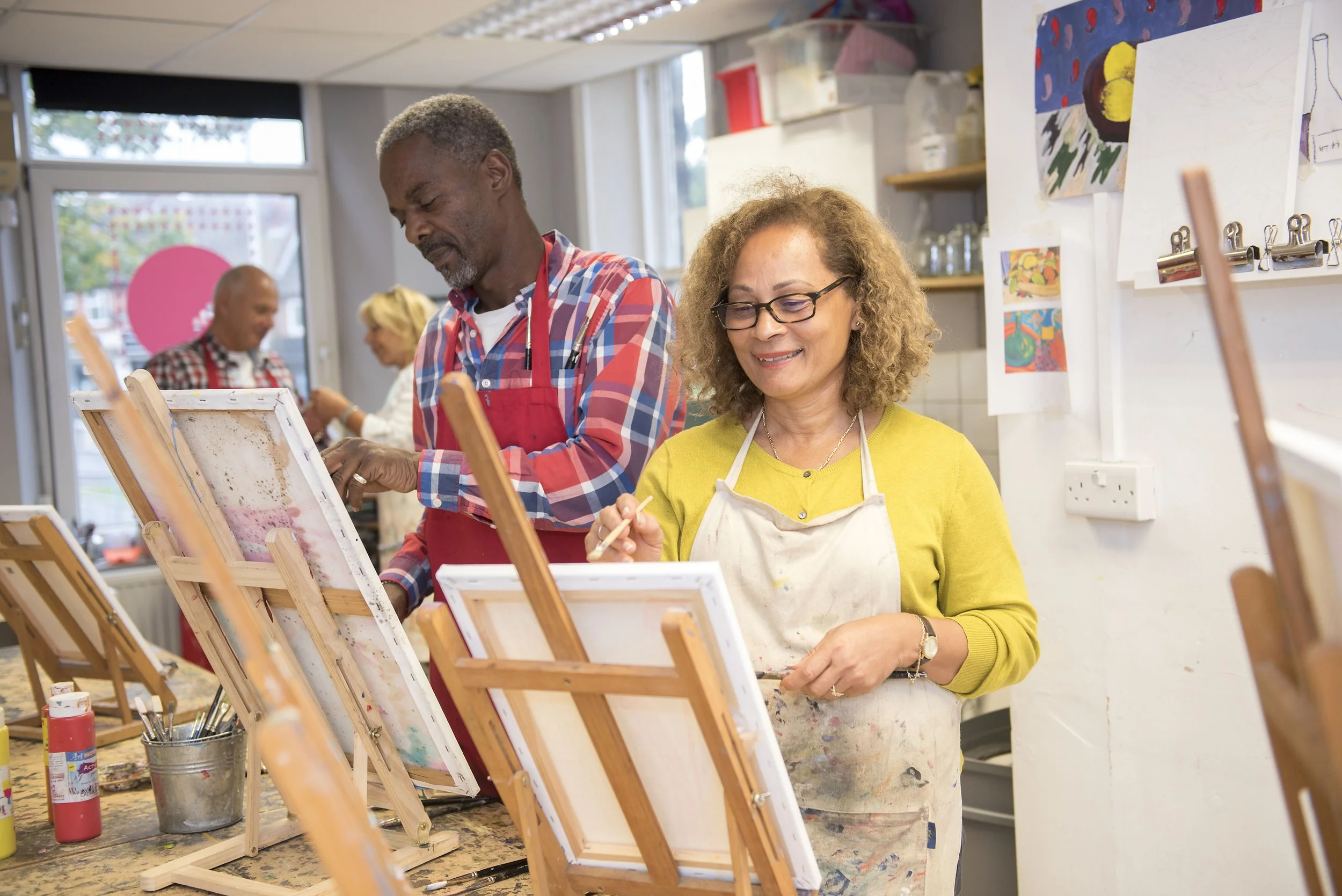 man and woman in an art class stood with easel, paint and brushes. he is in a check shirt and she in a light green top