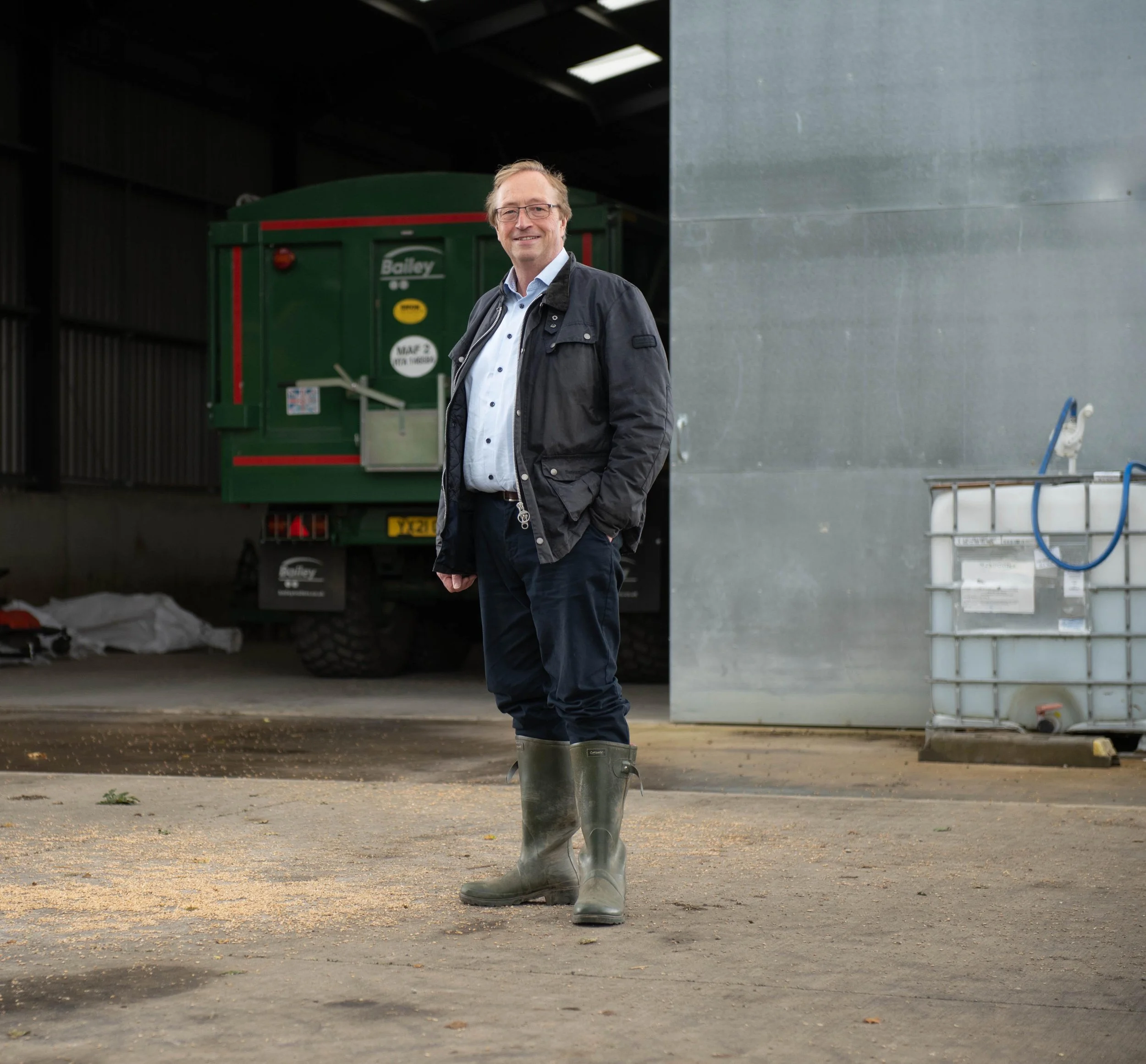 Man in a navy gilet standing in a farm building with a green truck and storage containers in the background, on-location business portrait