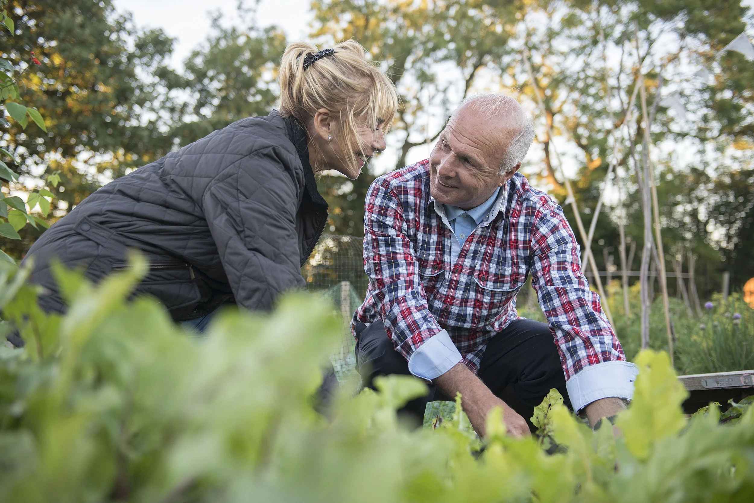 A woman and an elderly man smiling and talking while gardening outdoors, surrounded by green plants and trees in the background.