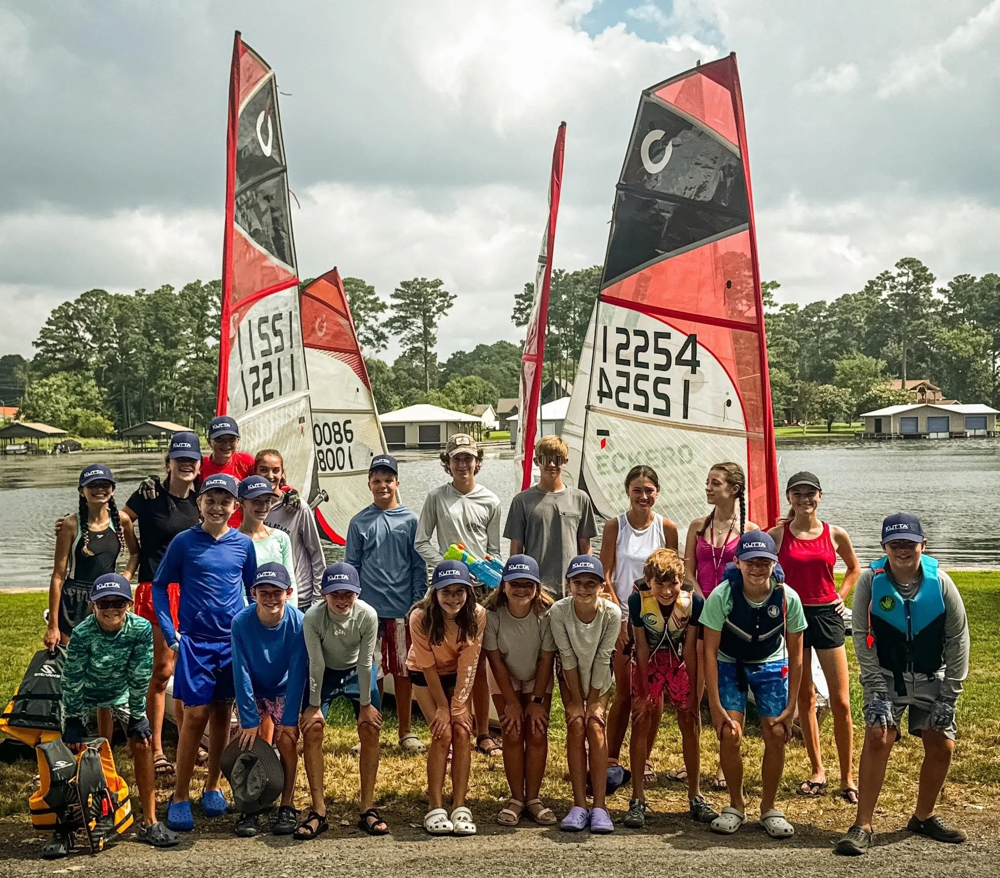 Group of children and teenagers smiling for a photo outdoors with sailboats and trees in the background.
