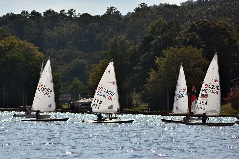 Four small sailboats with sailors, on a lake with sparkling water, surrounded by trees and hills.