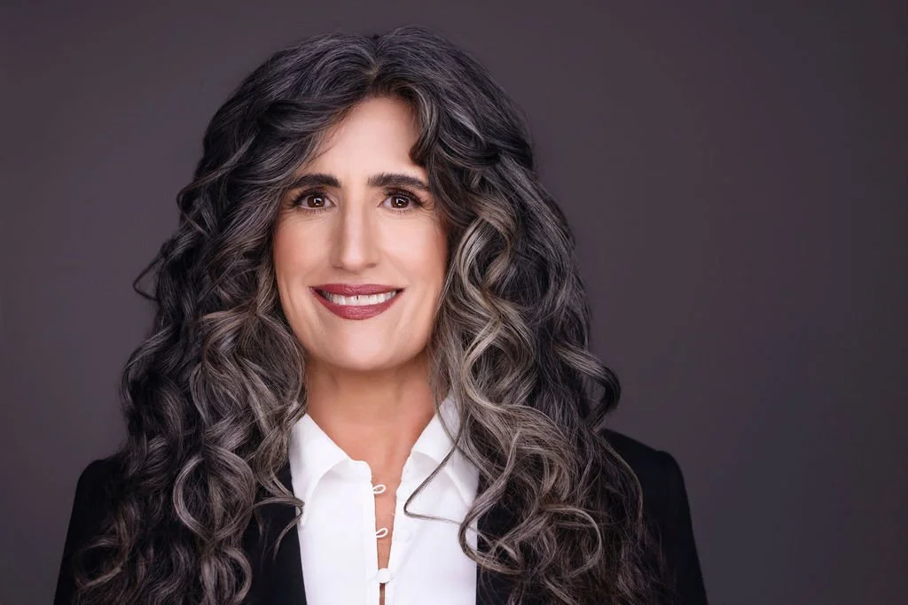 Smiling woman with long gray hair, in a white blouse, holding eyeglasses, seated in a chair with decorative pillows, interior room with framed quote on wall.