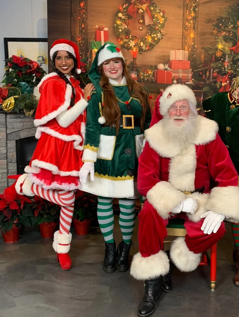 2 Elves and Santa Claus posing in Christmas costumes, with Christmas decorations in the background.