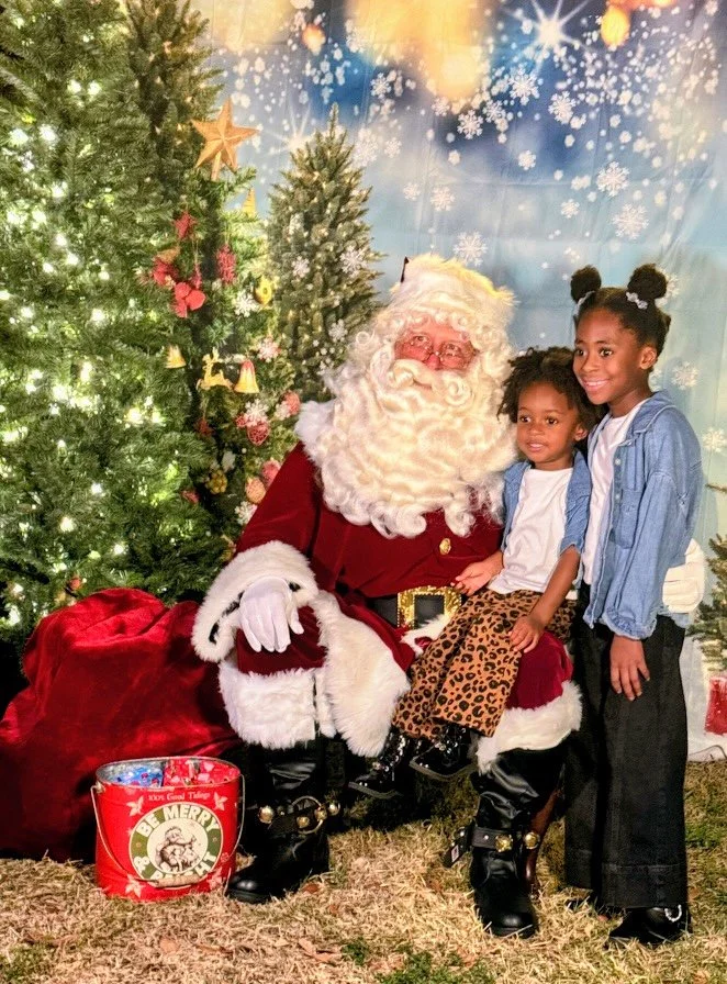 Two children with Santa Claus in Christmas setting with decorated trees and snowflake backdrop.
