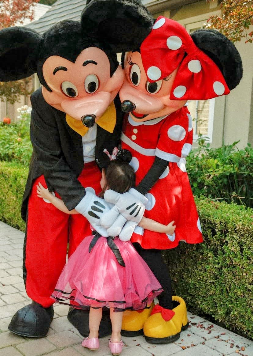 A young girl hugging costumed characters Mickey Mouse and Minnie Mouse outdoors on a sidewalk, with greenery and trees in the background.