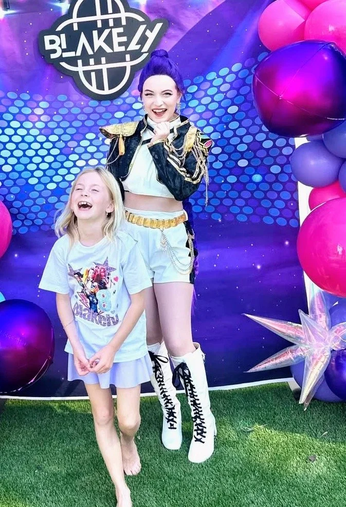 Rumi dressed in her iconic golden outfit with black jacket at a Dance Birthday Celebration smiling with the birthday girl laughing in front of her
