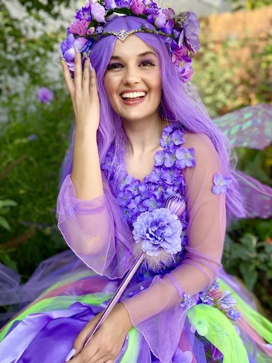 A young woman dressed as a fairy, wearing a purple flower crown, purple wig, and a purple dress adorned with flowers, smiling happily in a garden setting.