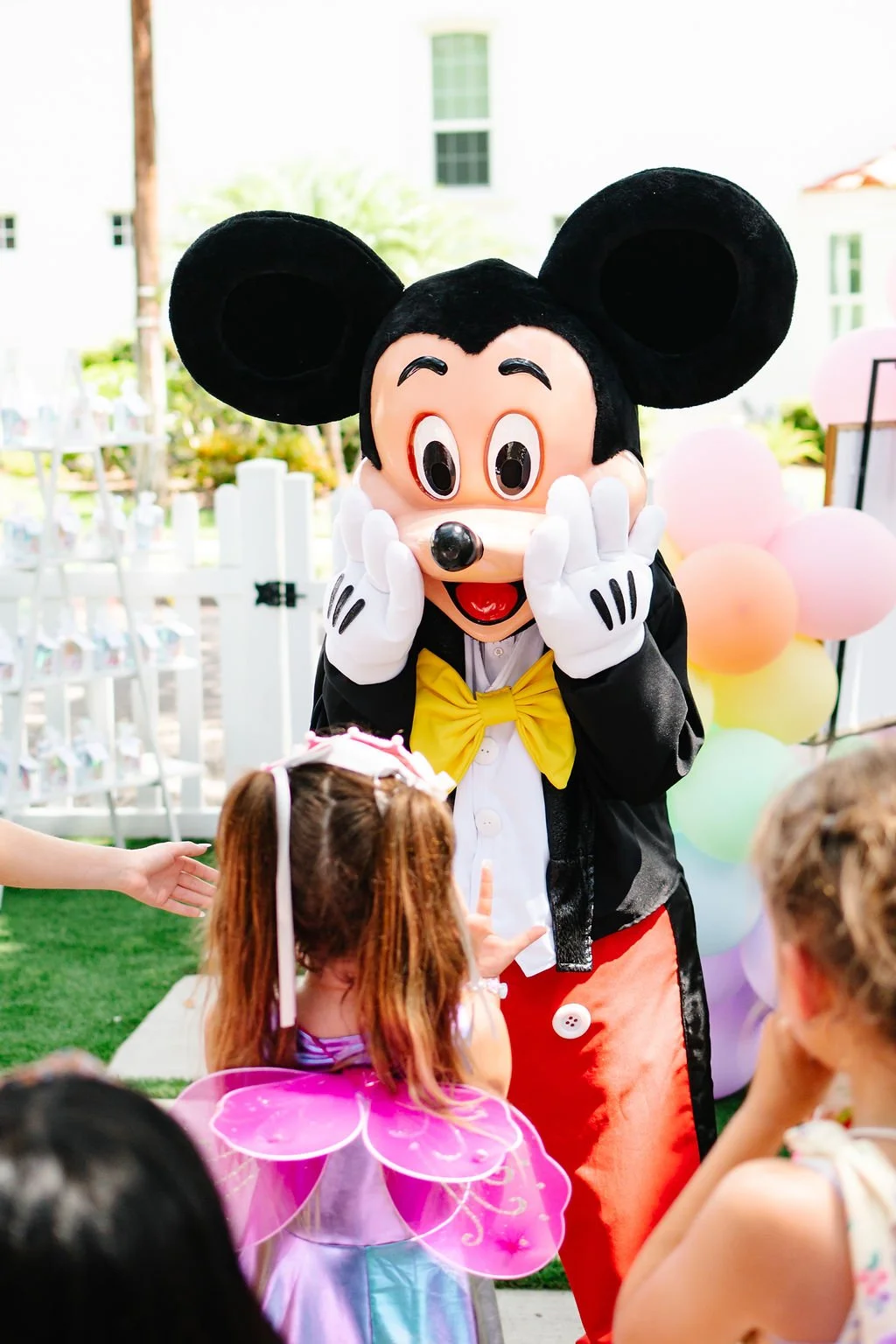 Mickey Mouse interacting with children at a celebration, with balloons in the background.