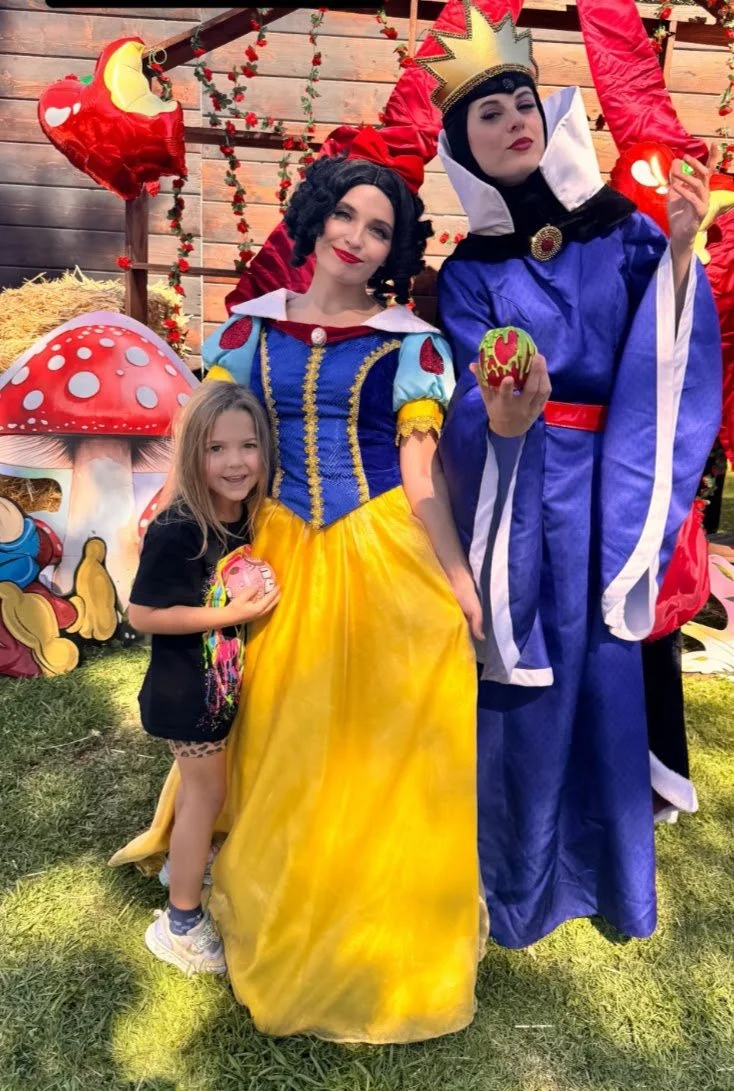 A young girl standing next to two women dressed as the Evil Queen and Snow White from fairy tales. The girl is holding a painted apple, smiling, and wearing a black T-shirt and animal-print shorts. 