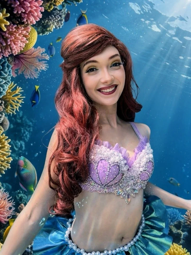 The Little Mermaid with long red hair smiling underwater, wearing a purple seashell-themed top decorated with pearls and a blue skirt, surrounded by colorful coral and fish.