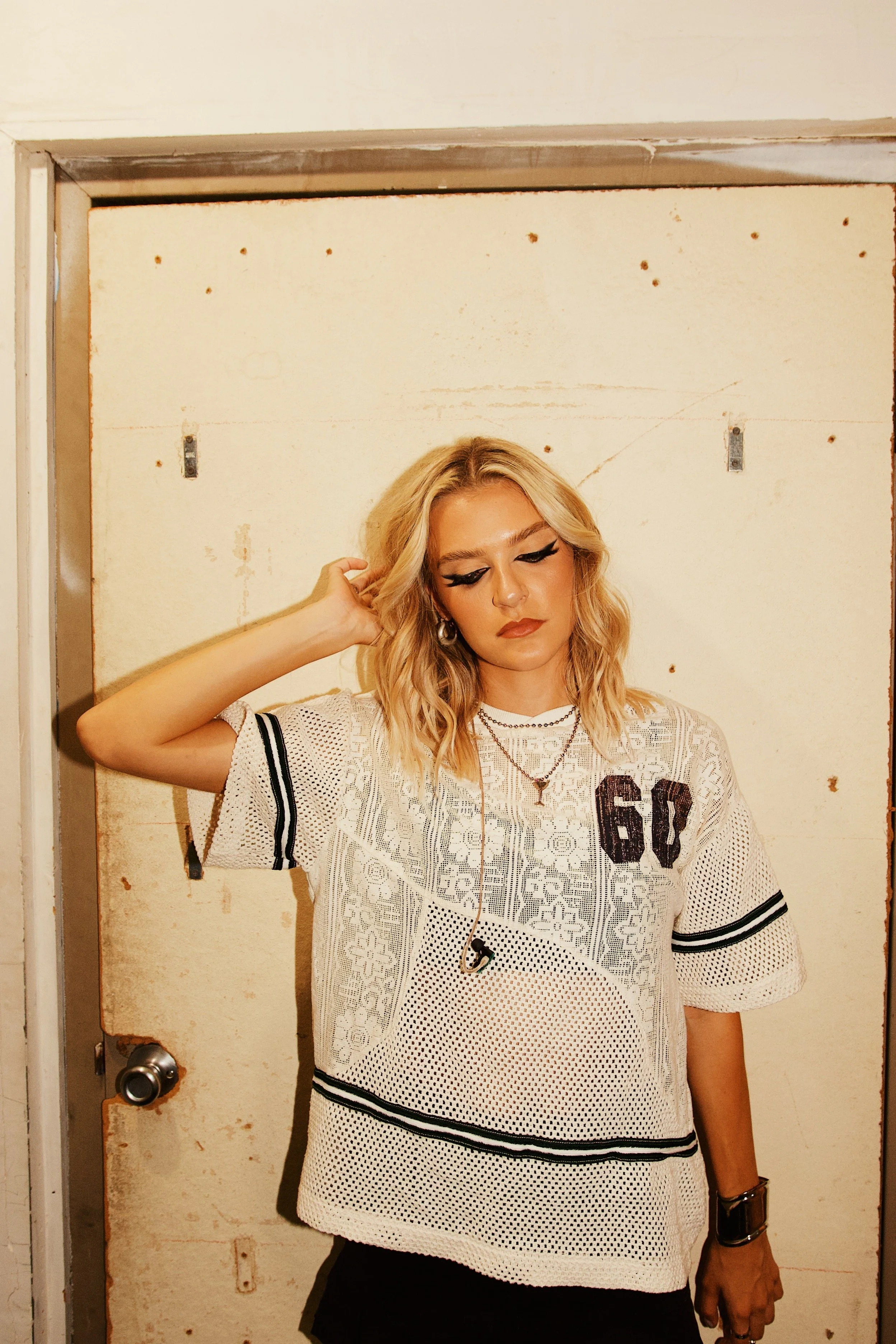 A woman with blonde hair, wearing makeup, a white lace short-sleeved dress with black stripes, layered necklaces, a black watch, and earrings, standing in front of a distressed beige door with rust spots and a door handle.