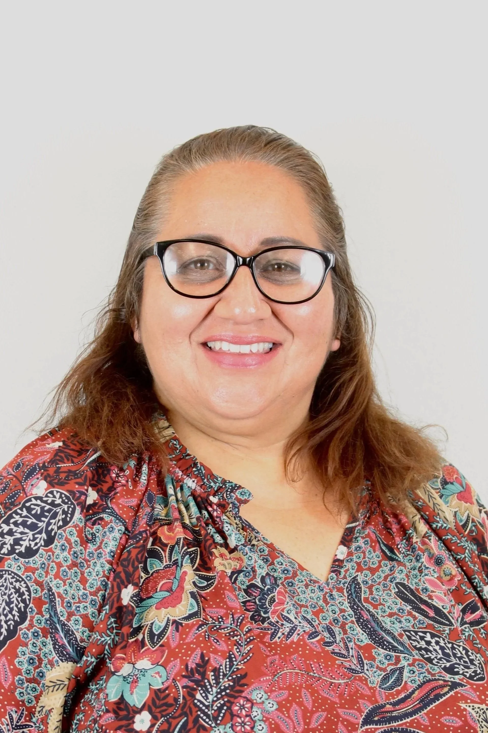A smiling woman with glasses and long brown hair, wearing a floral patterned blouse, against a plain light background.