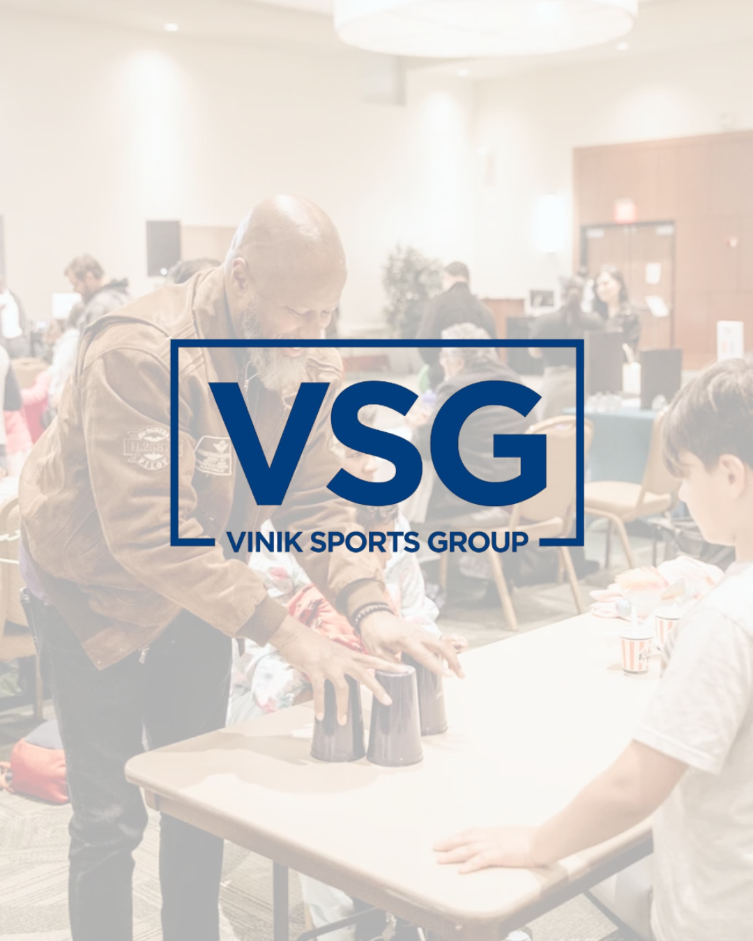 A man with a beard and a brown jacket performing a cup stacking activity with plastic cups at a table in a busy indoor event space, with people in the background. Overlay text says 'VSG VINIK SPORTS GROUP'.