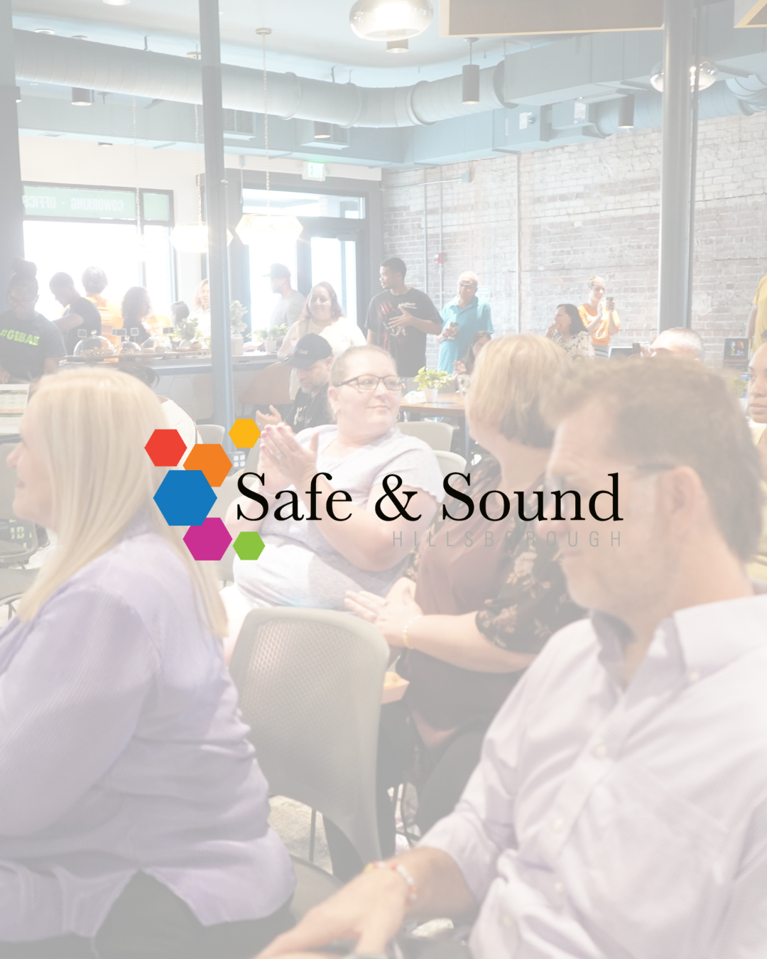 People gathered in a brightly lit, modern venue with exposed brick walls and industrial ceiling pipes, attending an event for 'Safe & Sound' in Hillsborough.
