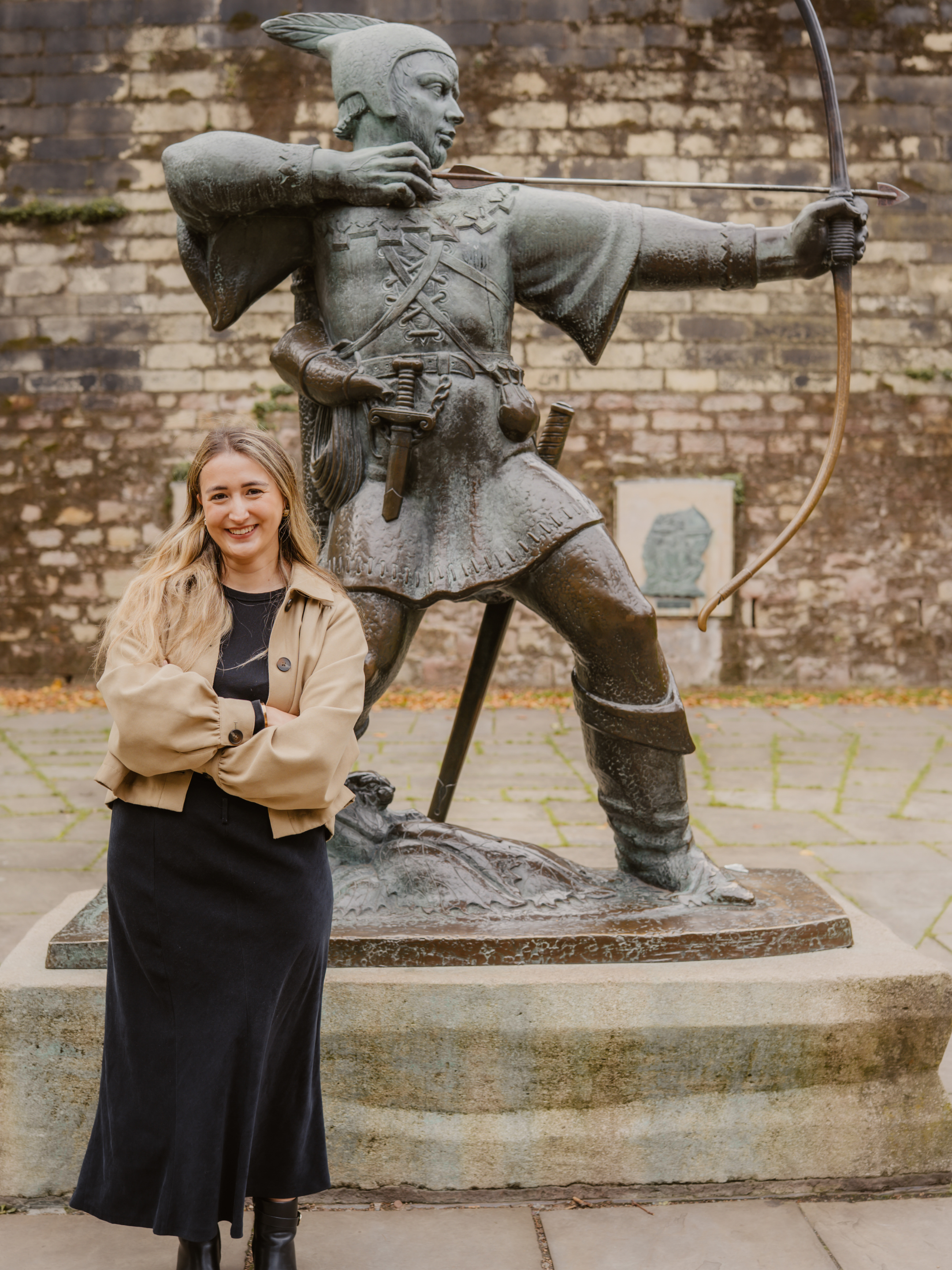 Eve Smallman standing in front of a Robin Hood statue wearing a black dress with her arms crossed, smiling