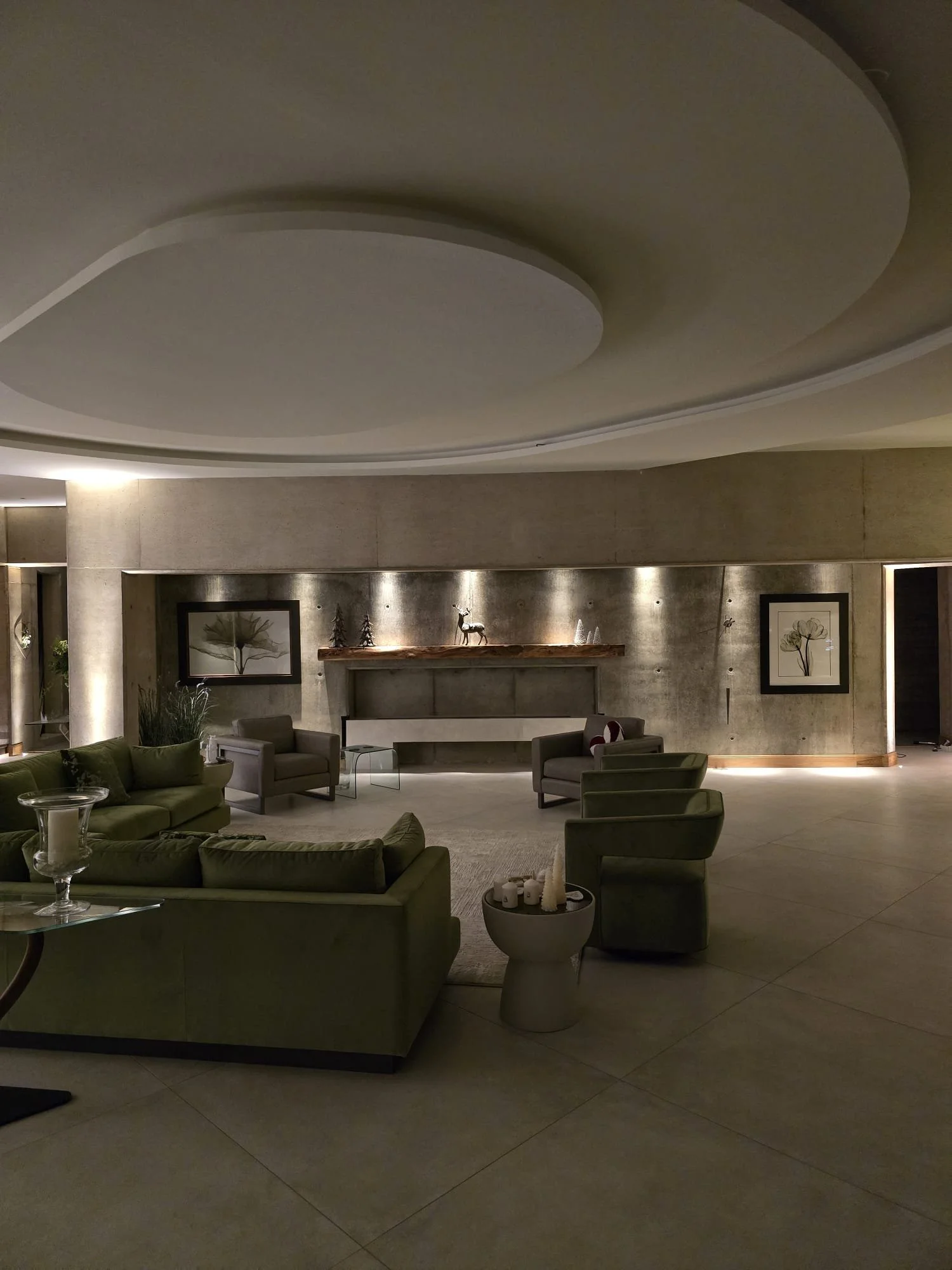 A modern hotel lobby with green sofas and armchairs, a concrete wall with framed artwork, and a decorative shelf with reindeer and trees, illuminated by soft lighting.