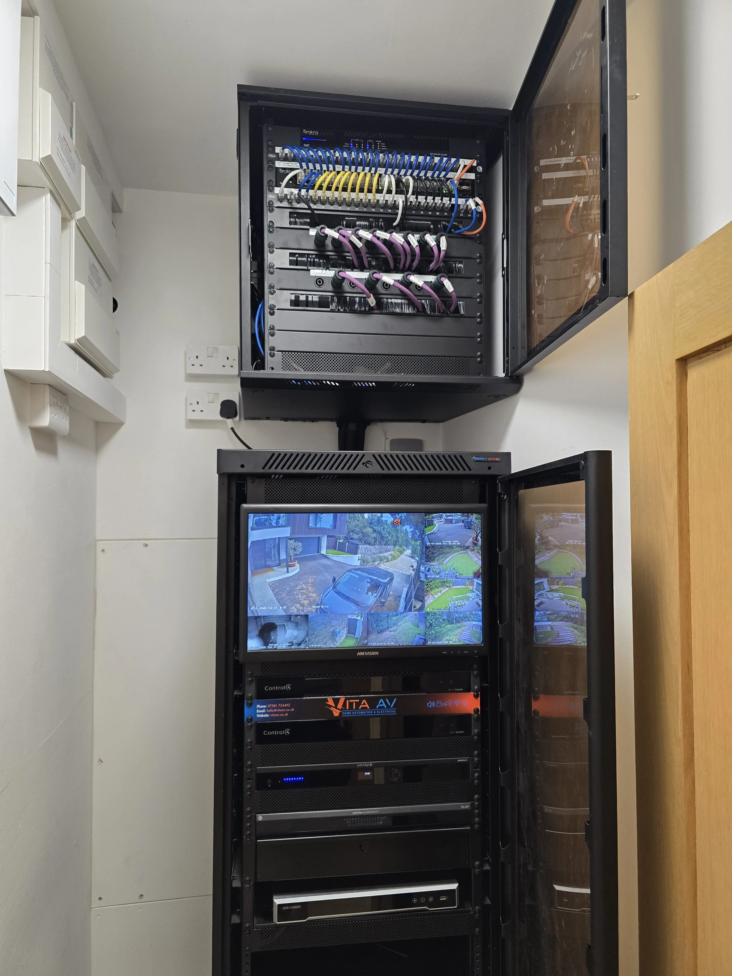 A server room with a tall black server rack housing various electronic equipment and a smaller black network cabinet with a glass door containing network switches and patch panels.