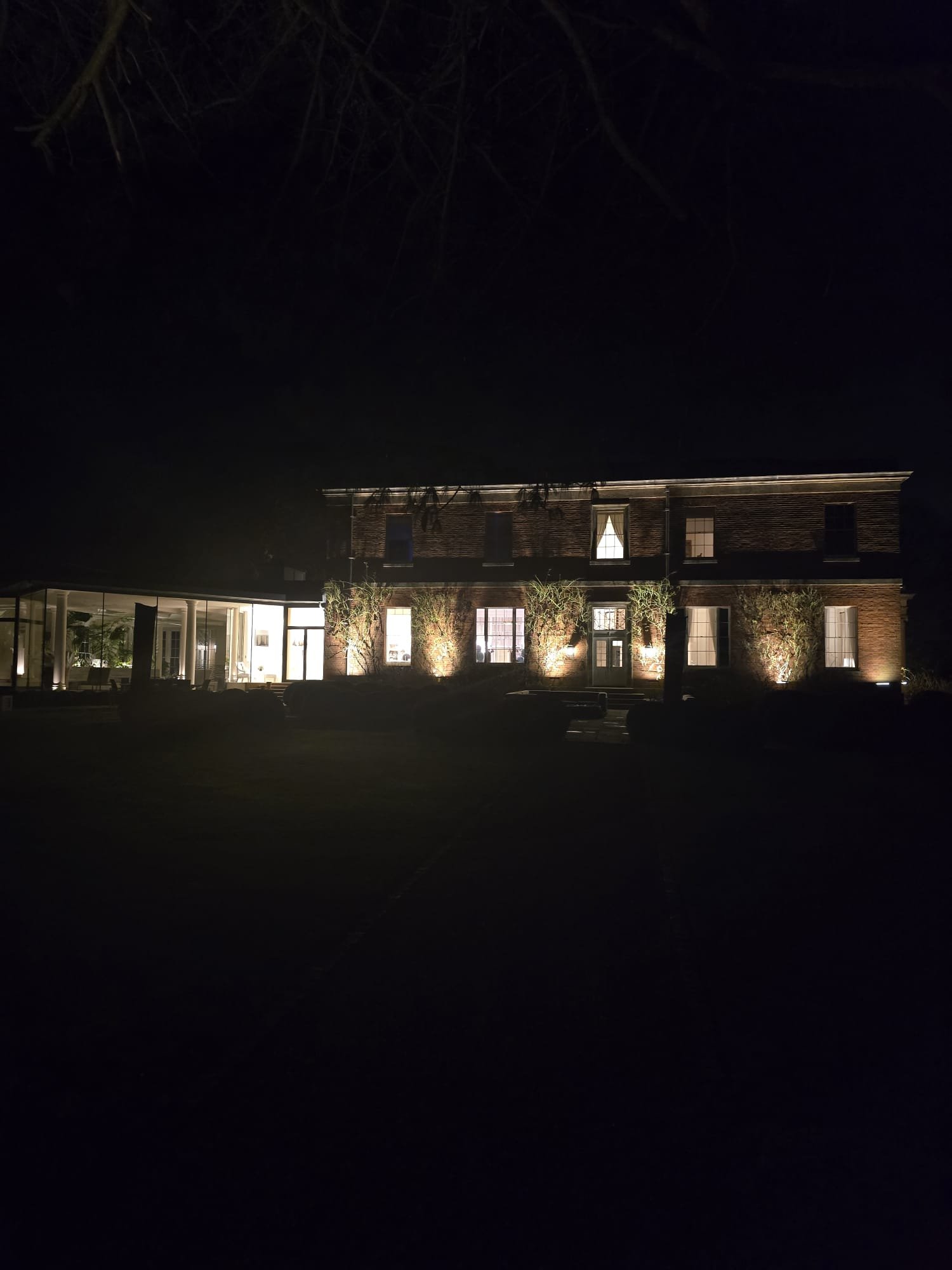 A large brick building illuminated at night with several lit windows and exterior lights highlighting trees and landscape in front.
