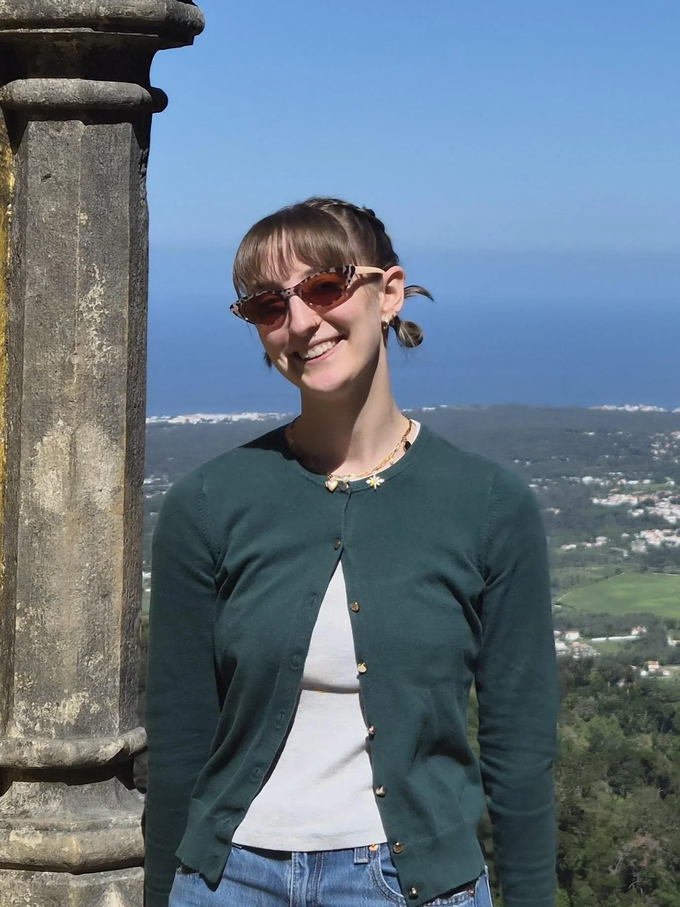 A woman smiling and wearing sunglasses, standing outdoors on a sunny day with a distant view of a valley and a large body of water in the background. She has short brown hair with a braid, a green cardigan, and a daisy necklace.