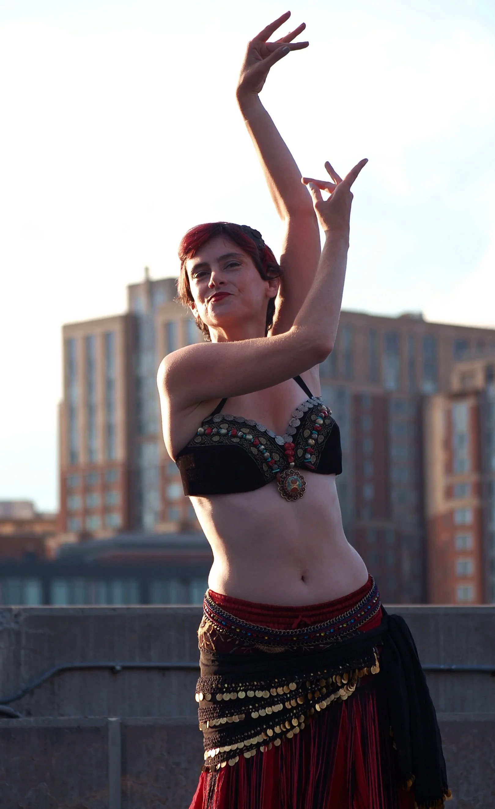 A woman with short red hair performing a dance outdoors against a cityscape backdrop, wearing a decorated top and a long black skirt with gold accents.