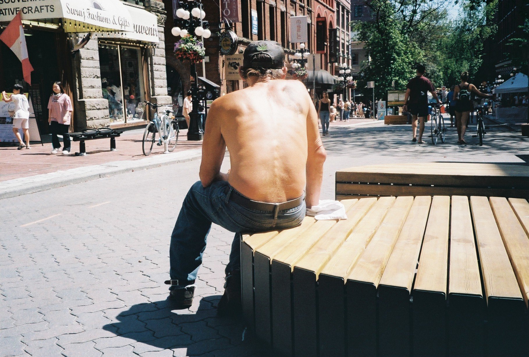 A shirtless man with a cap sitting on a bench in a city street, with shops, pedestrians, and bicycles in the background.