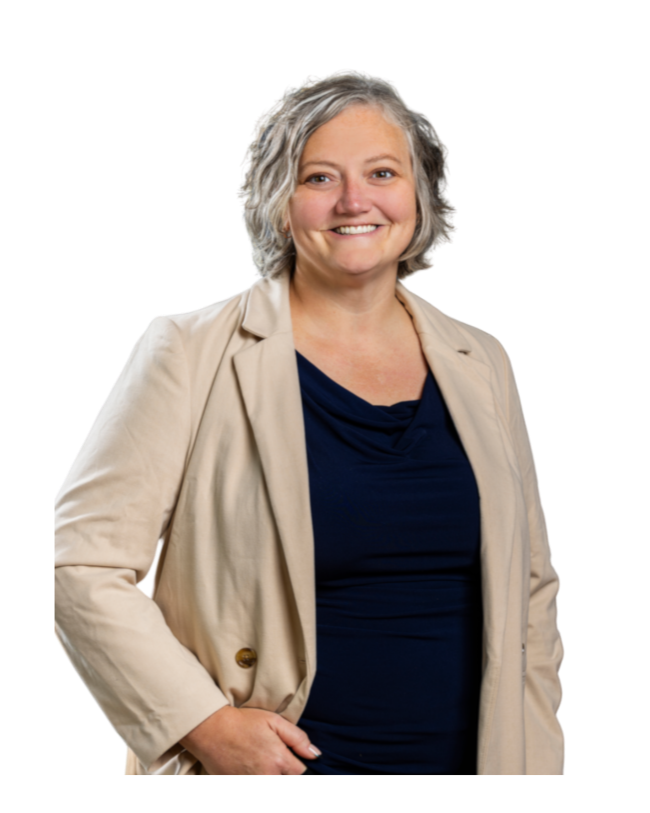 A smiling middle-aged woman with short, wavy gray hair, wearing a beige blazer over a navy blue top, standing with her right hand on her hip.