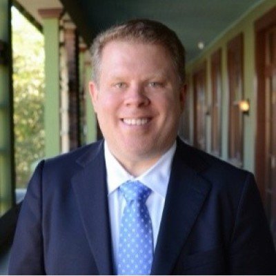 A smiling man in a navy suit and light blue tie standing indoors near a window with trees outside, with wooden doors and a hallway in the background.