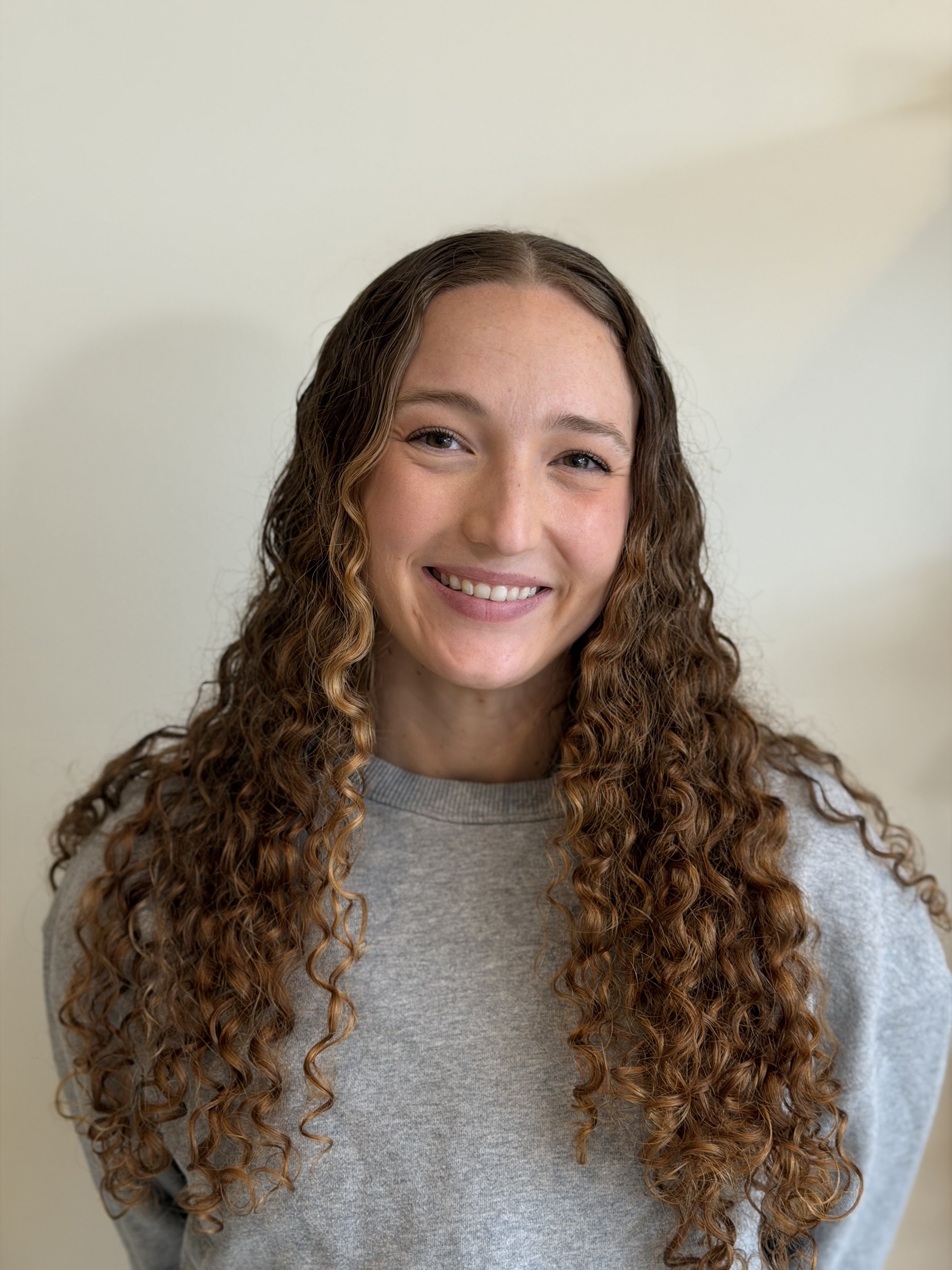a white women standing in front of a white wall with long curly red hair and a gray shirt