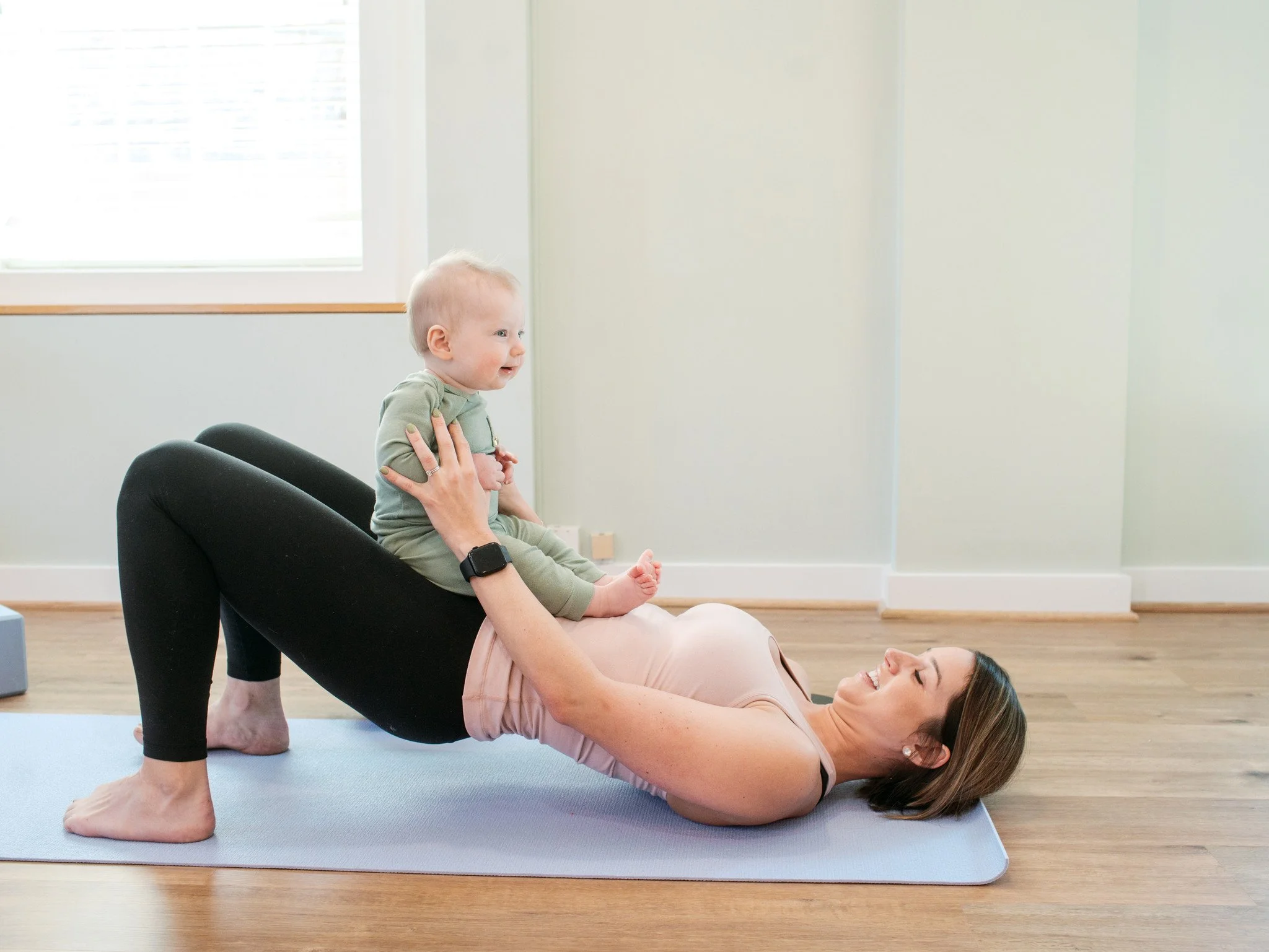 White woman with brown hair on a yoga mat doing a bridge with her baby on her belly