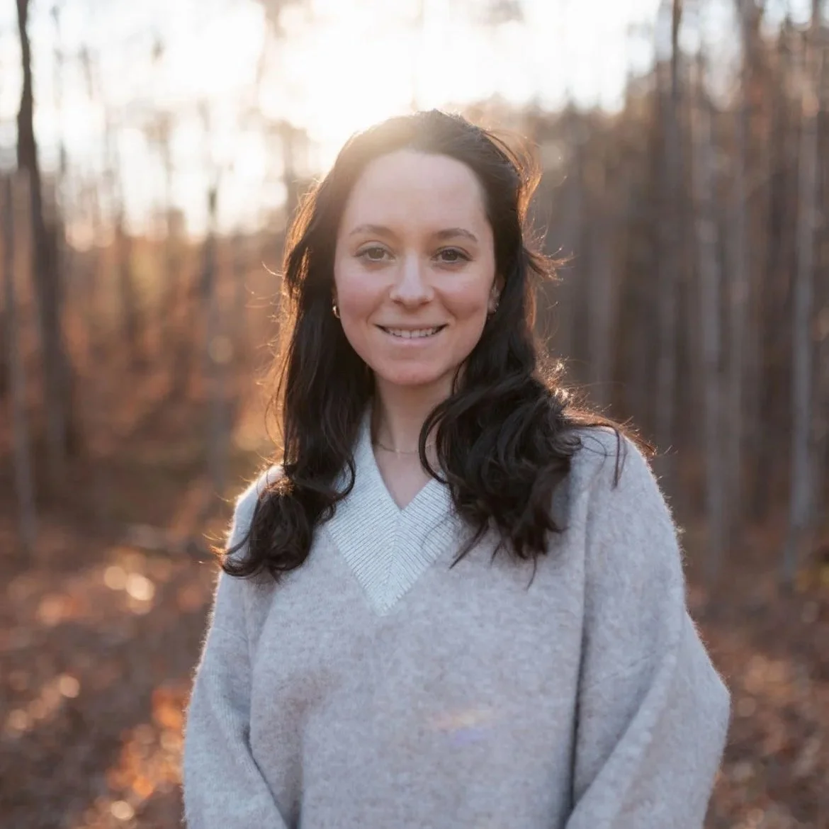 a profile of a white woman with brown hair, in a gray sweater with the sun setting behind her