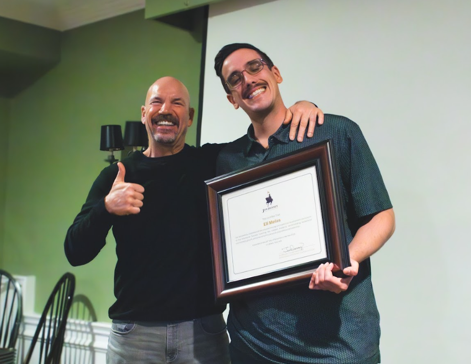 Two men smiling, one giving a thumbs up, the other holding a framed certificate.