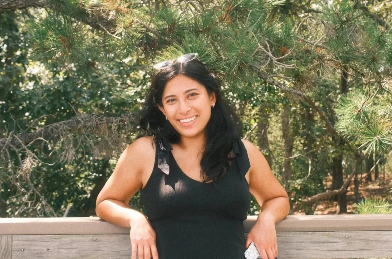 A young woman with long dark hair and a floral shirt standing outdoors, smiling at the camera during sunset with a background of trees and greenery.
