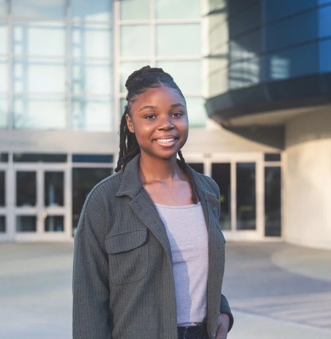 Young woman with braided hair and casual attire smiling outdoors in front of a modern glass building.