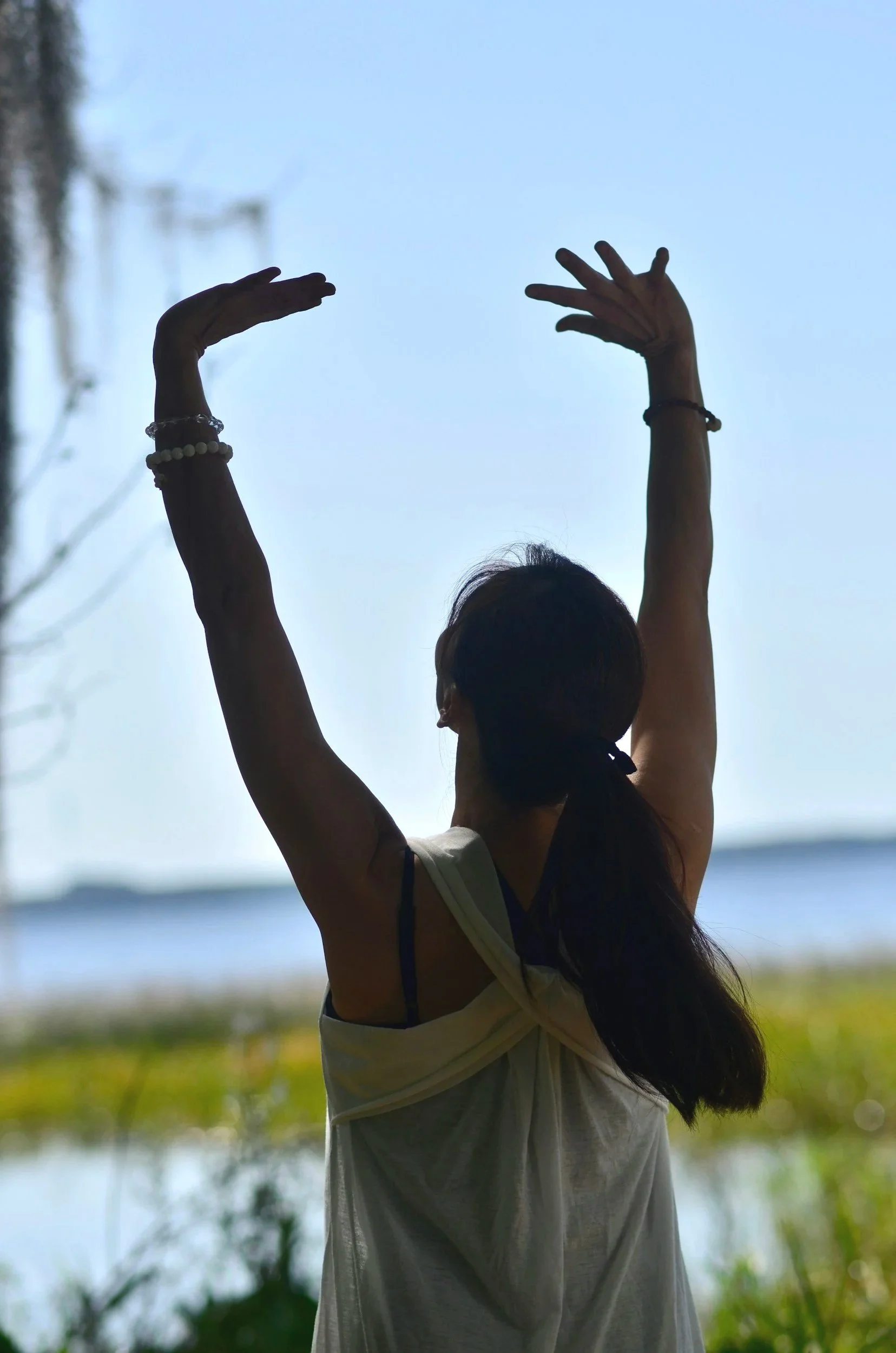 A woman standing outdoors near a body of water with her arms raised above her head, wearing a sleeveless top and bracelets, silhouetted against a bright sky.
