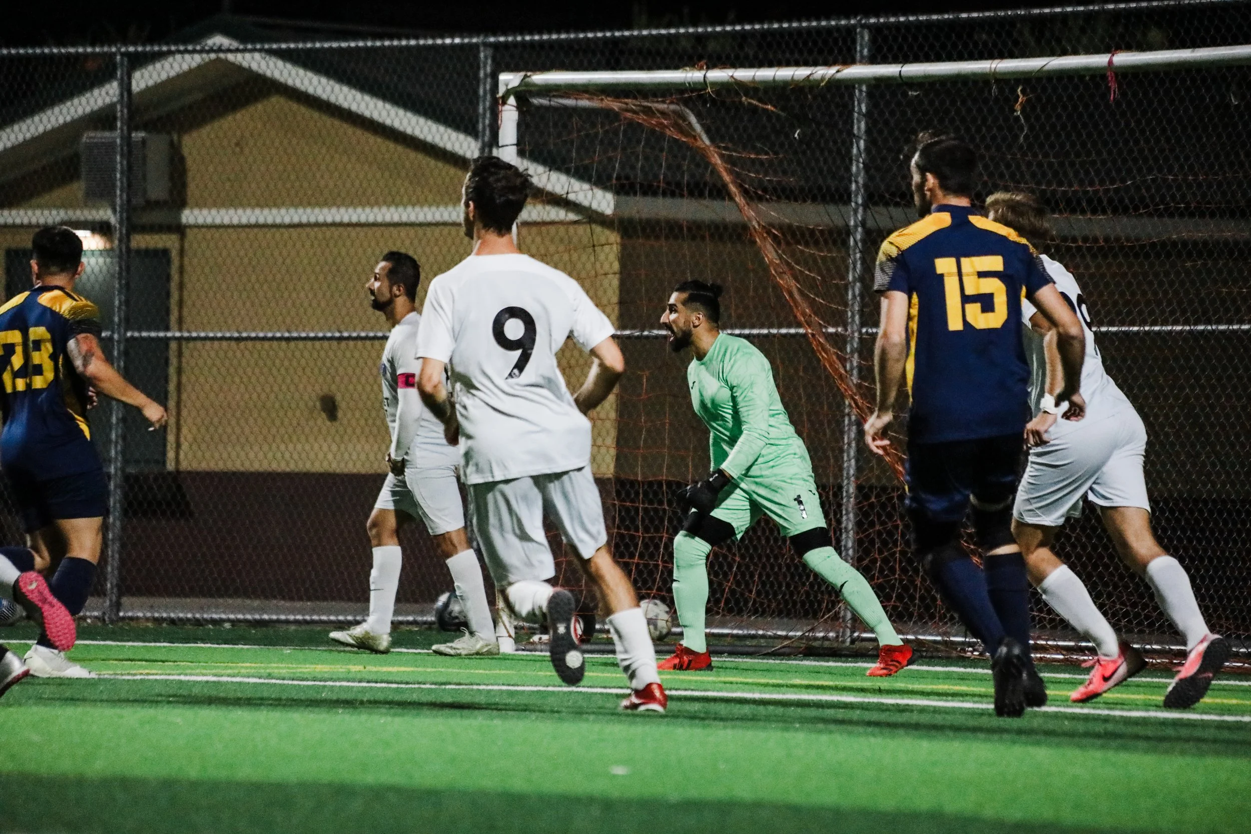 Soccer players on the field during a game at night. The scene includes a goalkeeper in a green kit preparing to defend the goal, surrounded by outfield players in white and dark blue uniforms. The match is taking place on a green artificial turf with a chain-link fence in the background.