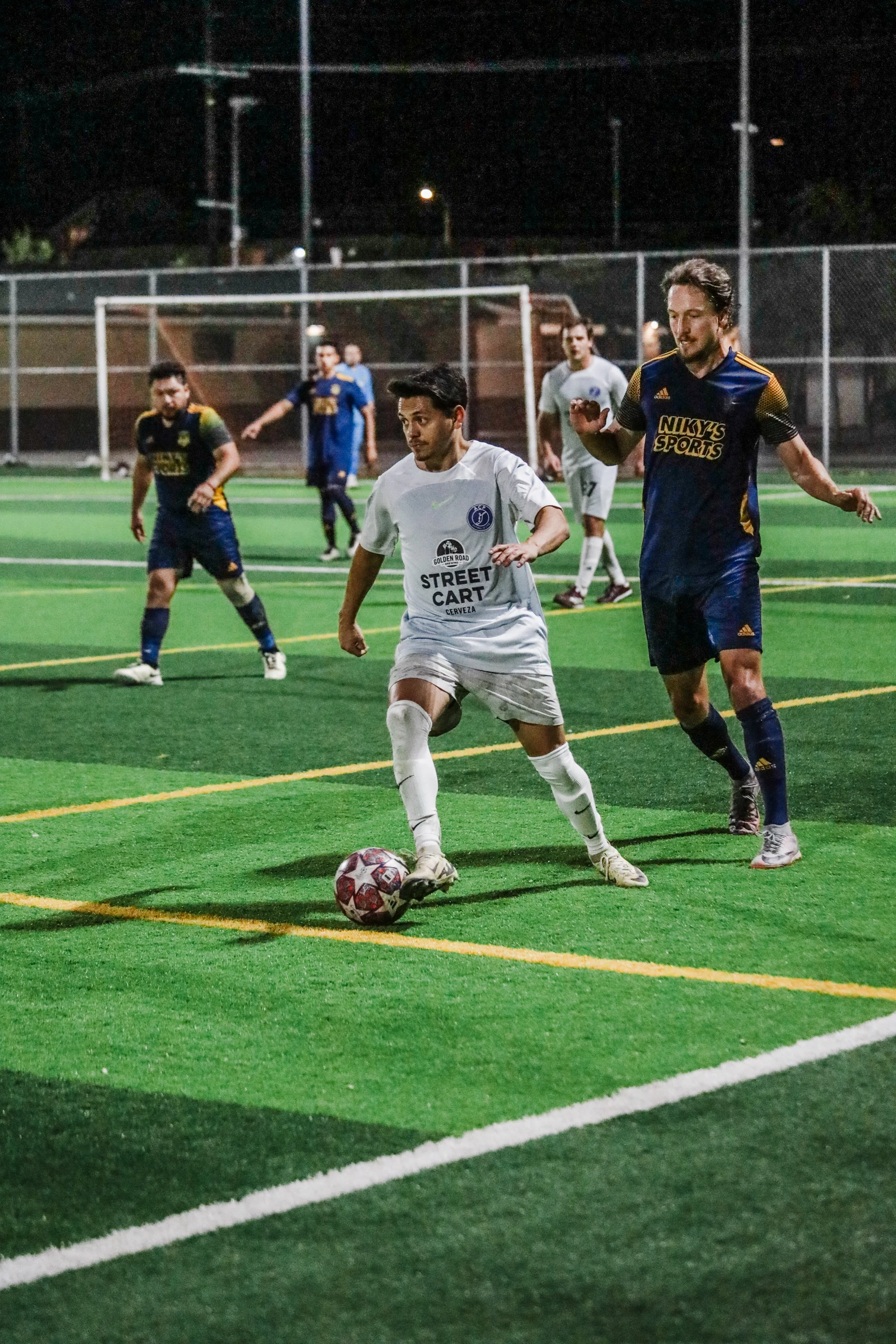 Soccer players competing for ball on night-time field, with one player in white controlling the ball while a player in blue and gold attempts to block.