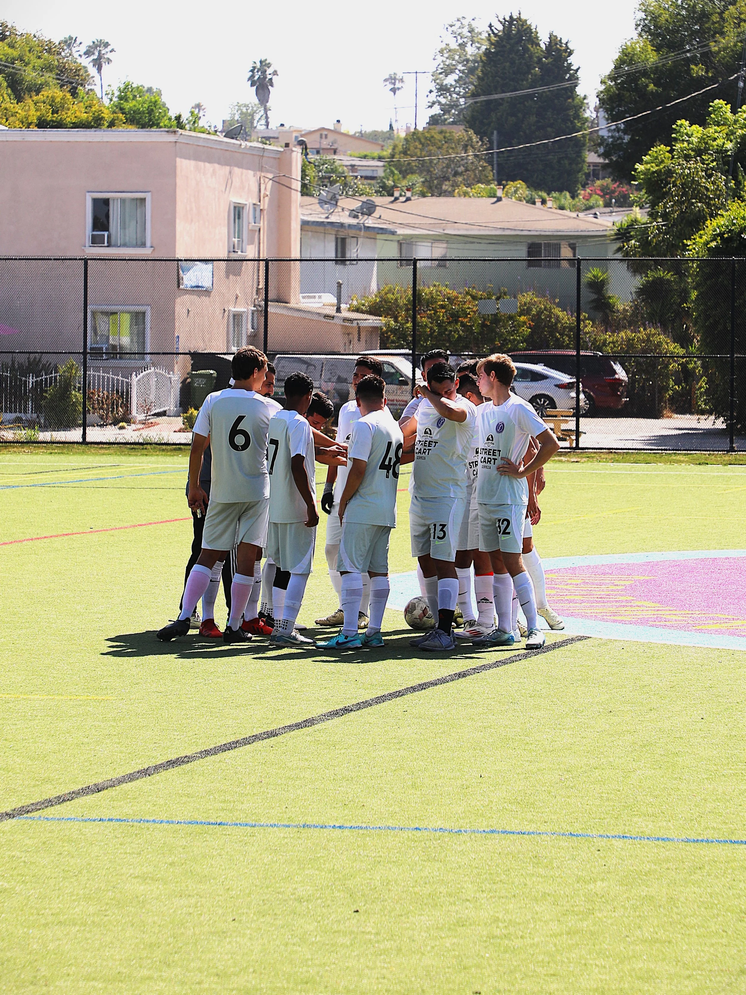 A group of young soccer players in white uniforms huddling on a field during daytime, with a fence, residential buildings, and trees in the background.
