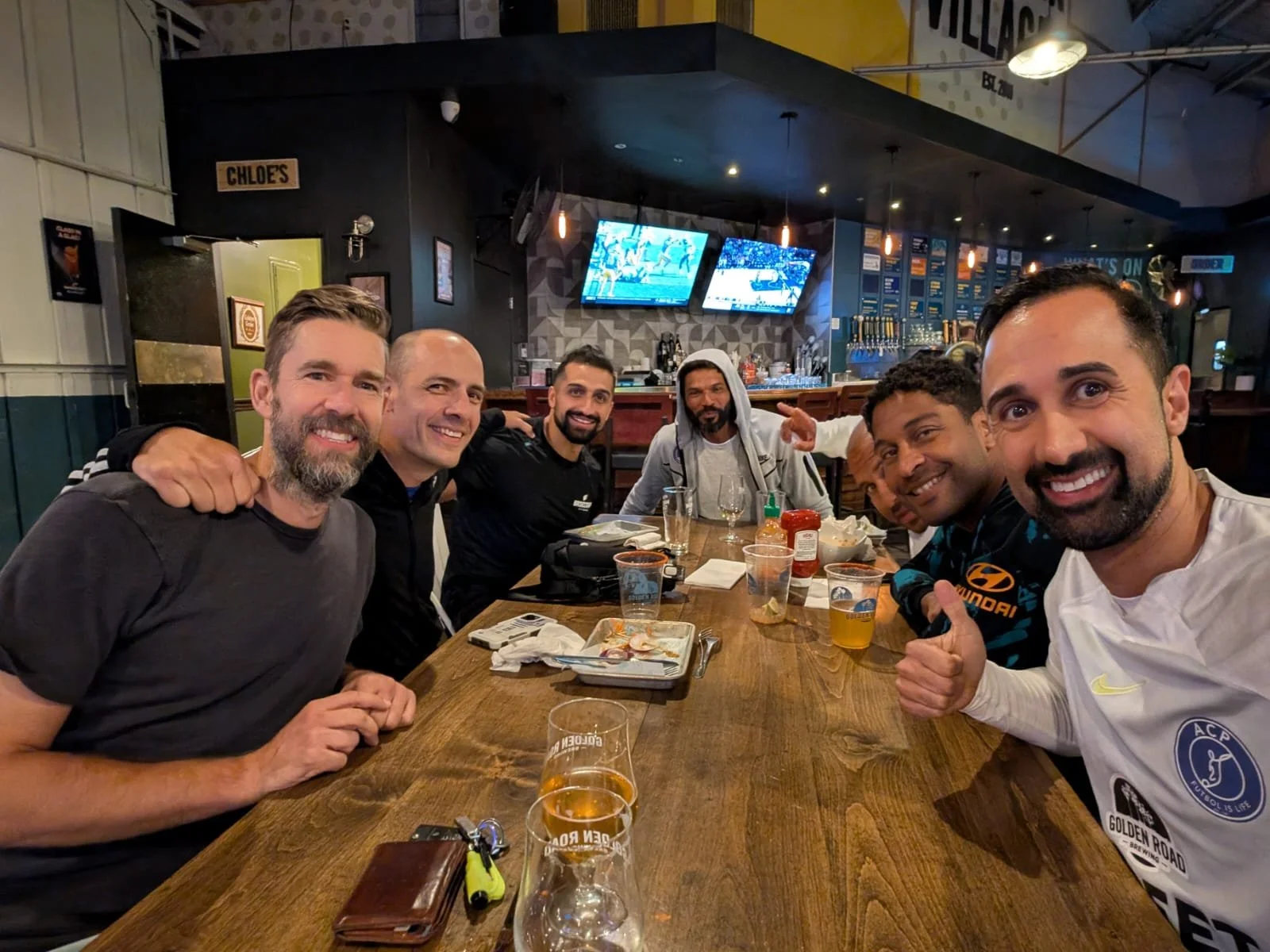 A group of seven men sitting around a wooden table in a bar or restaurant, smiling and enjoying drinks. There are multiple TVs on the wall and a bar area in the background.