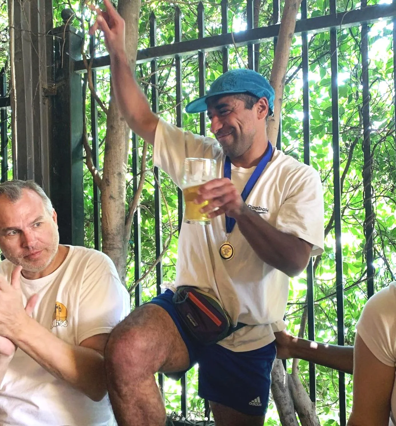 A man in a white t-shirt and blue shorts is holding a beer and smiling while sitting on a fence with one leg raised. He is wearing a blue cap, a medal around his neck, and a small waist pouch. There are other people nearby, with a man on the left looking at him, in an outdoor setting with green foliage in the background.