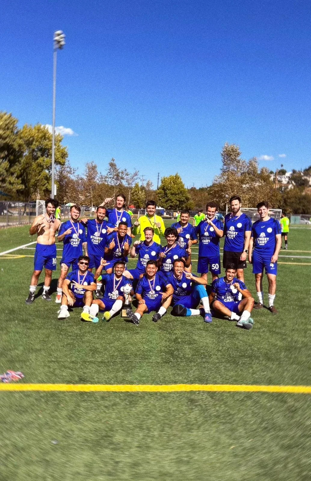 A group of soccer players in blue uniforms celebrating on a field after a game, most wearing medals around their necks.