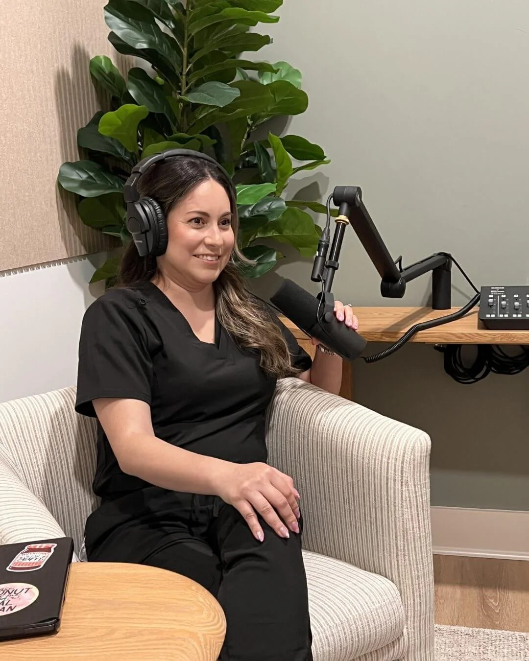 A woman with long brown hair wearing black scrubs, sitting on a beige striped armchair, smiling, wearing large black headphones, and holding a microphone positioned in front of her mouth in a recording studio or podcast setup. There is a large green plant behind her, a wooden table with a laptop with stickers on it, and a desk with audio equipment.