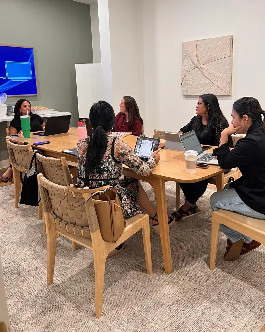 A business meeting with five women sitting around a wooden table in a modern conference room, each using a laptop or tablet, with drinks on the table, a television on the wall, and minimalistic wall art.