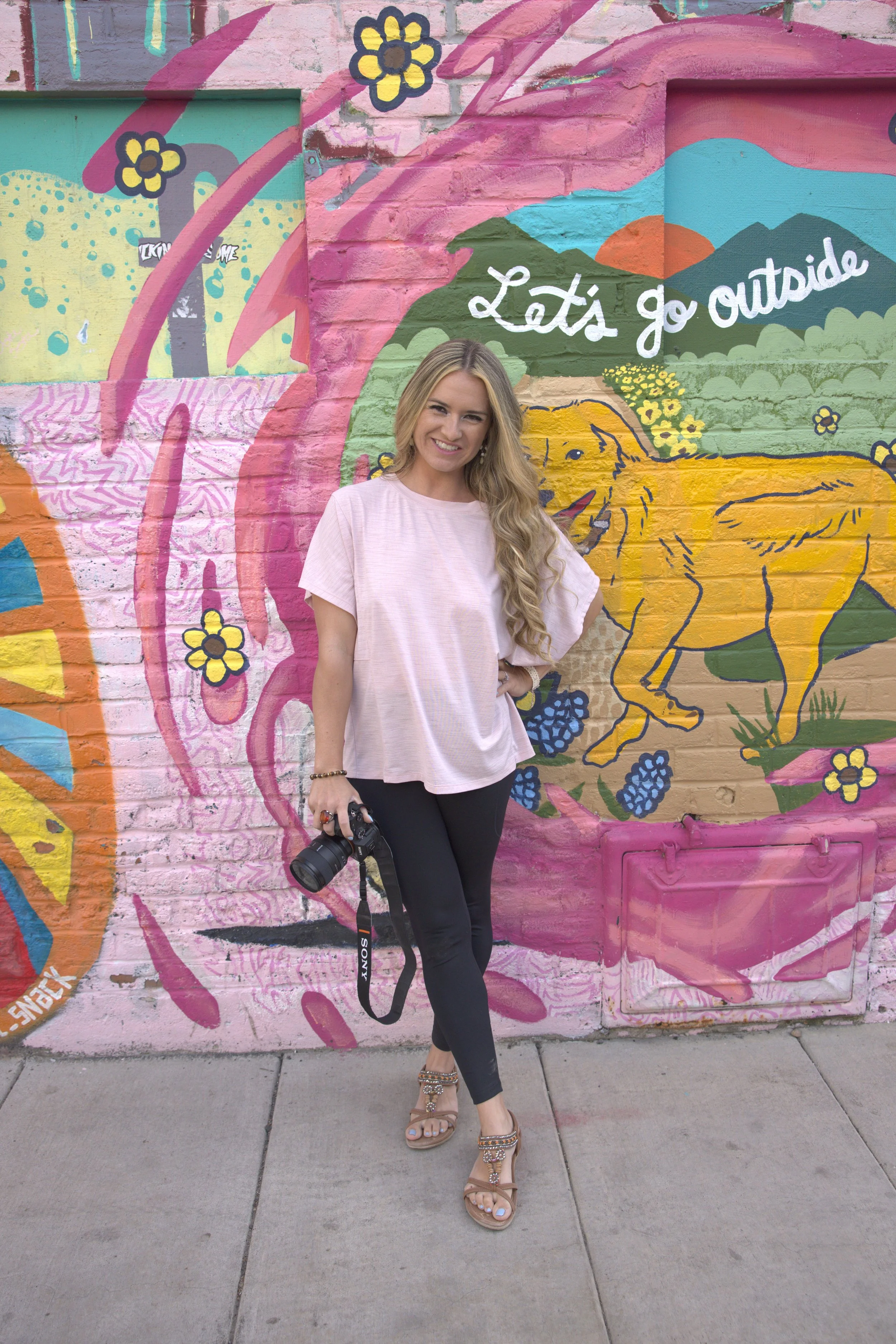 Colorado photographer holding a camera, posing in front of a bright mural with mountains, flowers, and the phrase “Let’s go outside.”