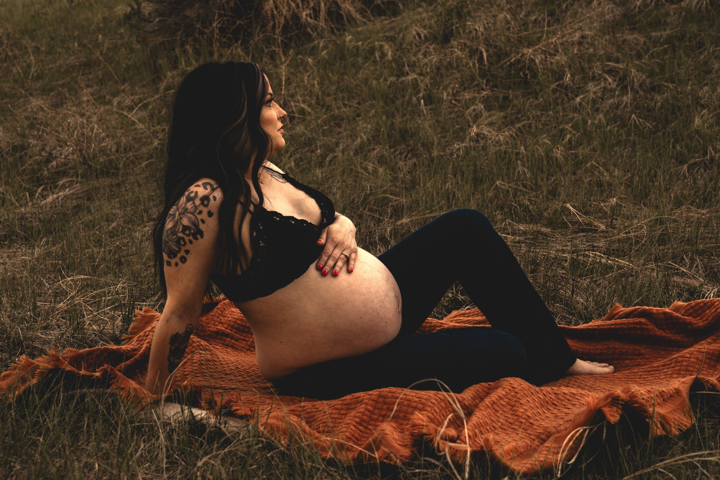 Expecting mom sitting on a blanket staring off pondering while resting her hand at the top of her belly.